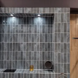 Narożna wnęka wyłożona pionowymi, ciemnymi płytkami z jasną fugą, oświetlona punktowymi lampkami, z dekoracyjnym szklanym pojemnikiem i małym okrągłym lusterkiem na półce, sąsiadująca...