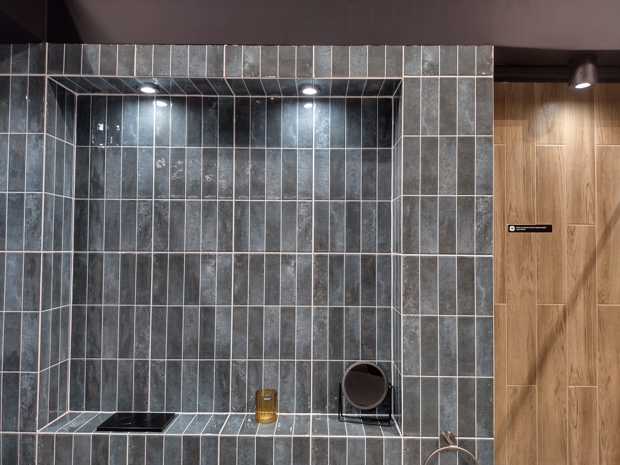 Narożna wnęka wyłożona pionowymi, ciemnymi płytkami z jasną fugą, oświetlona punktowymi lampkami, z dekoracyjnym szklanym pojemnikiem i małym okrągłym lusterkiem na półce, sąsiadująca...