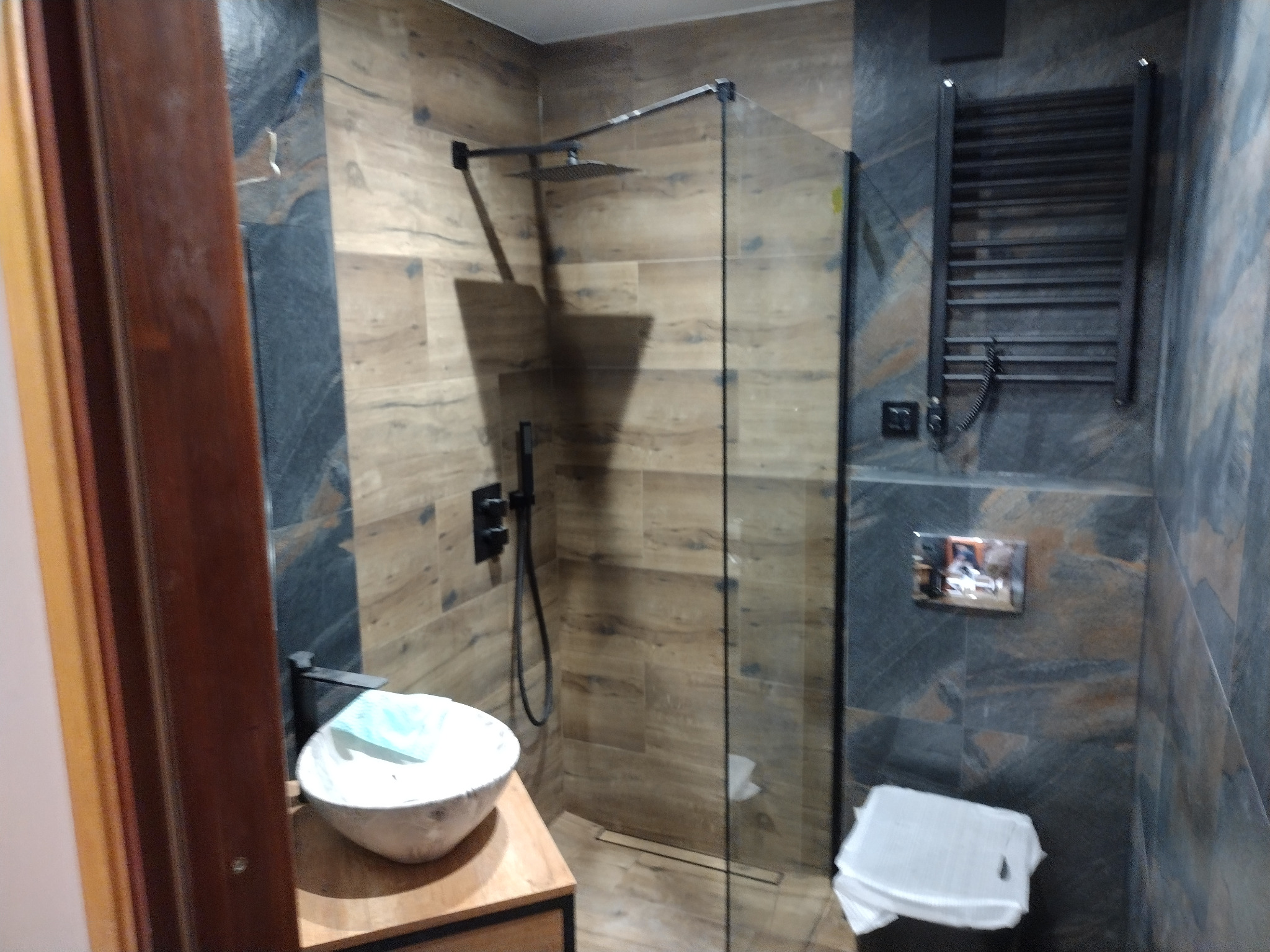 Nowoczesna łazienka z kabiną prysznicową typu walk-in, umywalką nablatową na drewnianym blacie, grzejnikiem drabinkowym i płytkami imitującymi drewno oraz szary kamień.