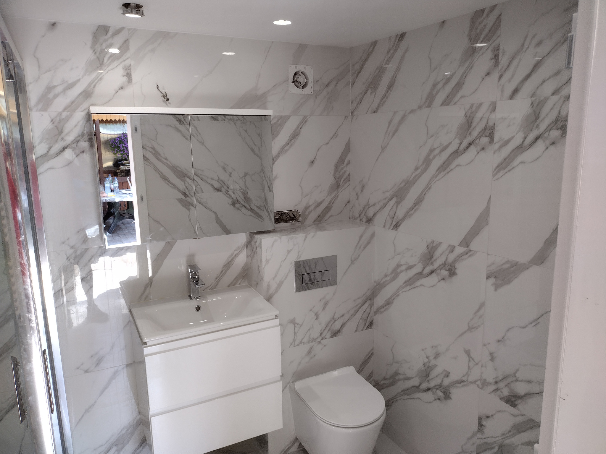 Nowoczesna łazienka z marmurowymi płytkami, wiszącą toaletą i szafką z umywalką, widoczny fragment kabiny prysznicowej po lewej stronie.