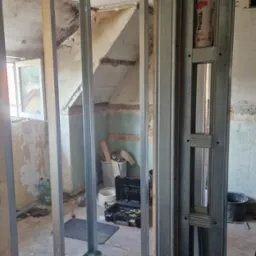 Stelaż z profili stalowych do ścianki działowej z płyty karton-gips w trakcie montażu w remontowanym pomieszczeniu ze starą powłoką malarską, widoczny otwarty kufer z narzędziami i butelka Fruvita...