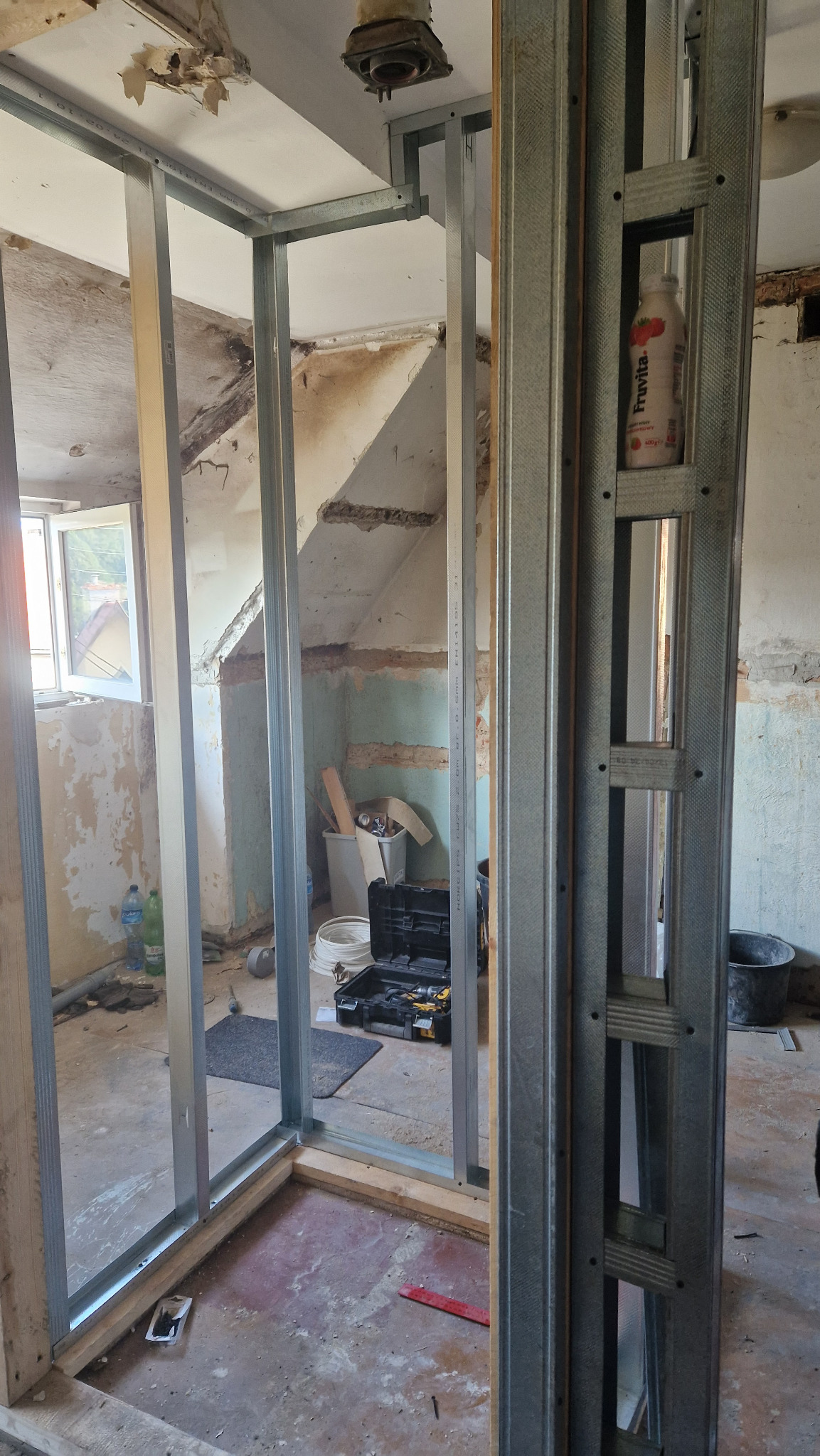 Stelaż z profili stalowych do ścianki działowej z płyty karton-gips w trakcie montażu w remontowanym pomieszczeniu ze starą powłoką malarską, widoczny otwarty kufer z narzędziami i butelka Fruvita...