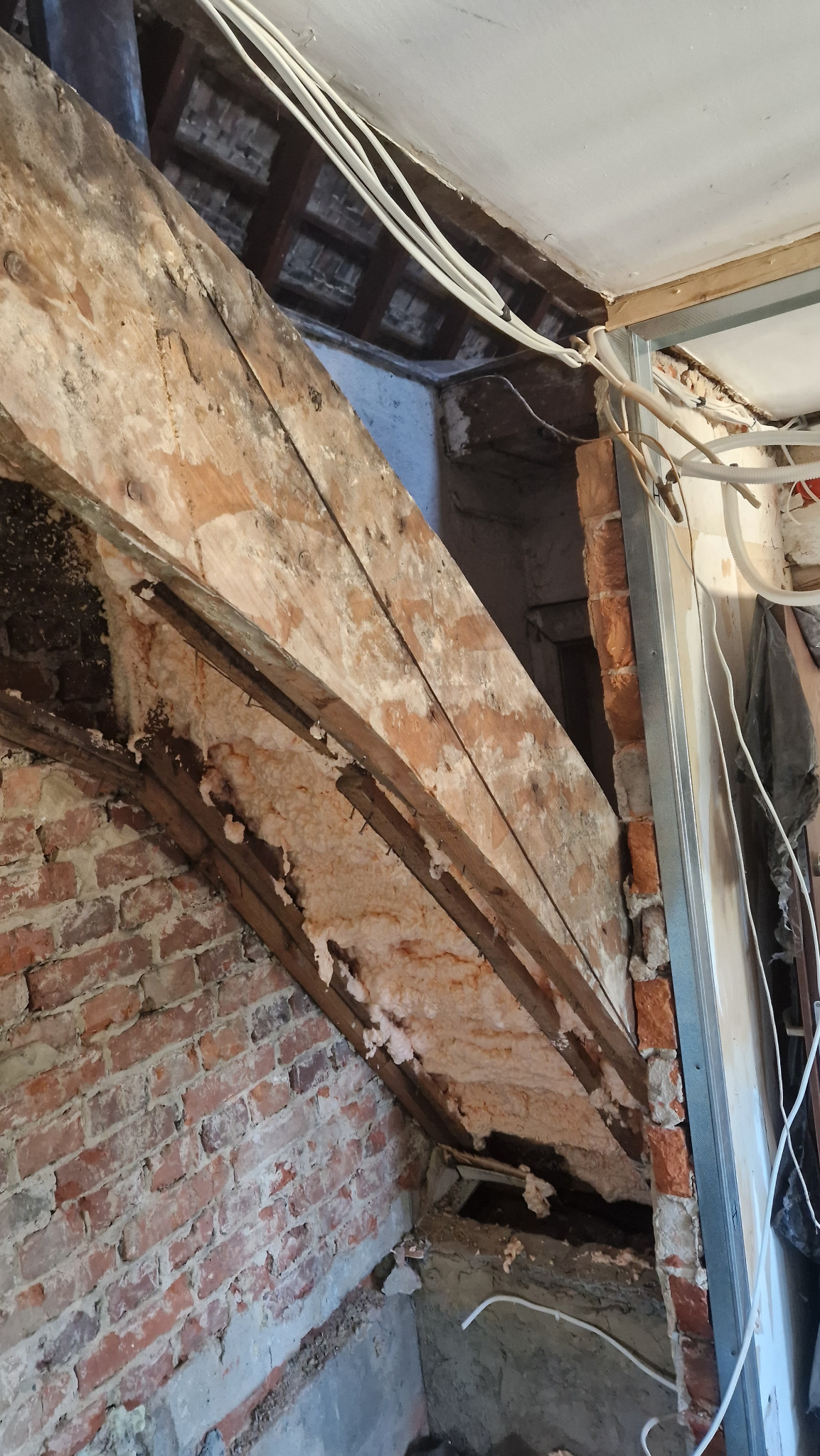 Ściana z czerwonej cegły z drewnianą konstrukcją i fragmentami pianki izolacyjnej, widoczne kable elektryczne i metalowy profil ściany działowej w trakcie prac.