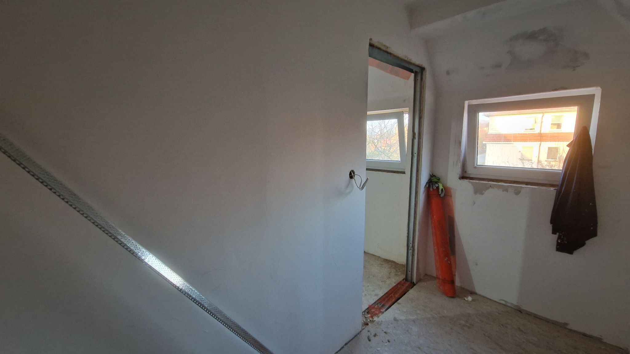 Wnętrze w trakcie remontu: ściana po gładzi z profilem metalowym, widoczne okna, przewody elektryczne i resztki materiałów budowlanych.