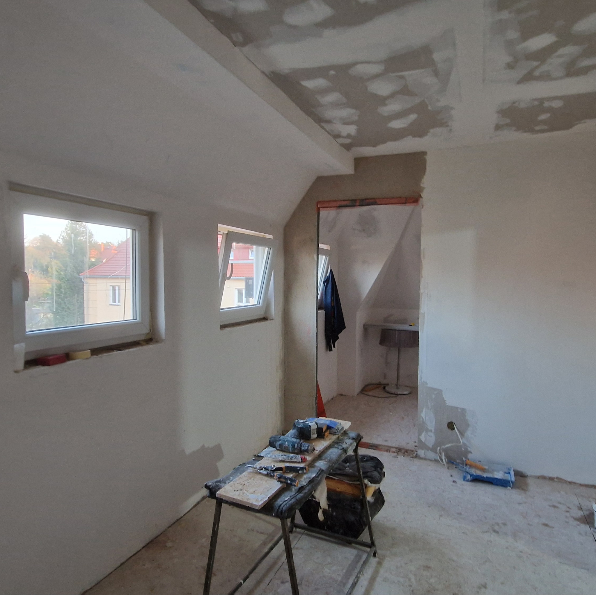 Poddasze w trakcie prac wykończeniowych, widoczne okna dachowe, częściowo wykończona ściana i sufit z płyt kartonowo-gipsowych, narzędzia na kozłach malarskich, w tle widoczne pomieszczenie z lampą.