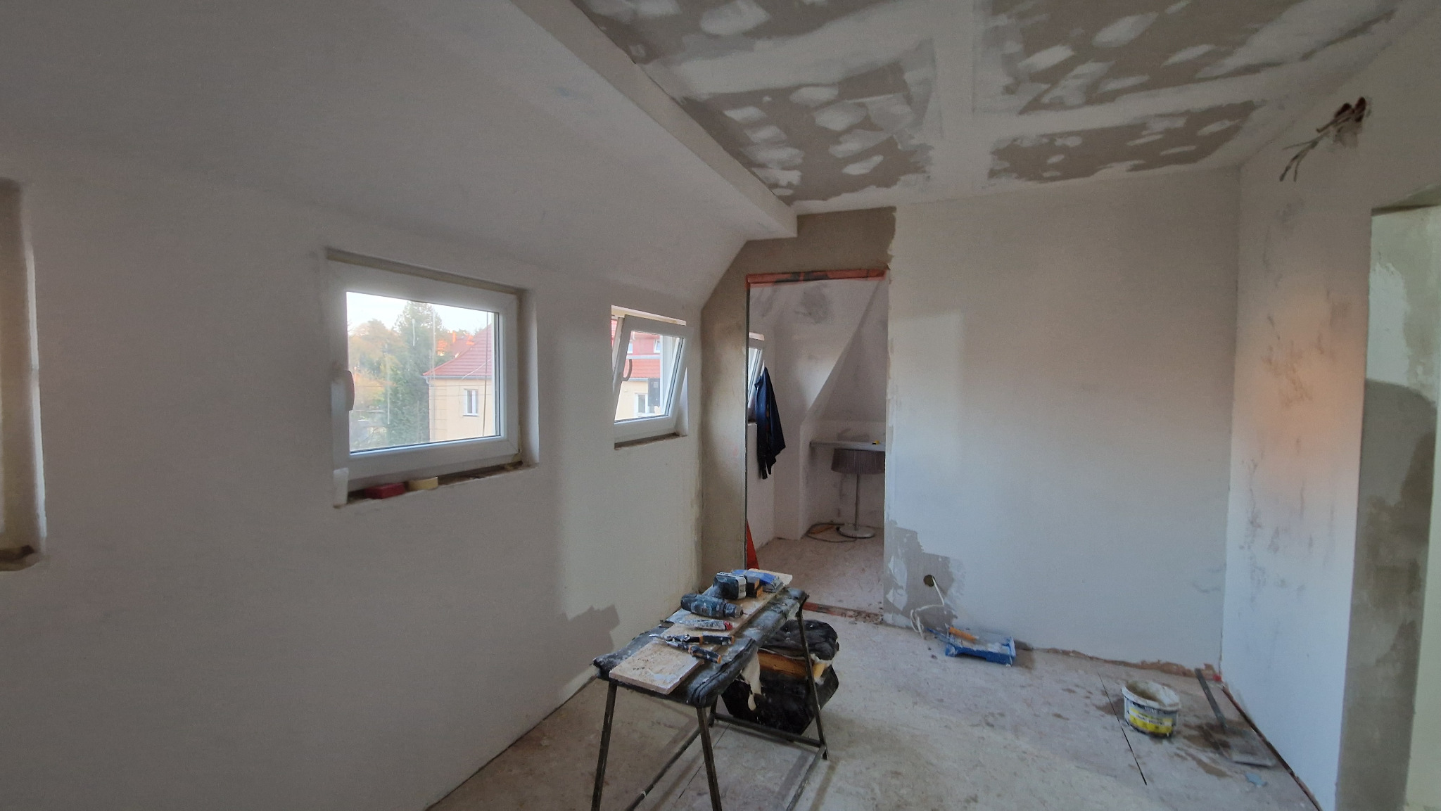 Poddasze w trakcie prac: gładzie na ścianach i suficie, widoczne narzędzia na stole warsztatowym, dwa okna dachowe z widokiem na zewnątrz, przygotowane ściany do dalszych prac.