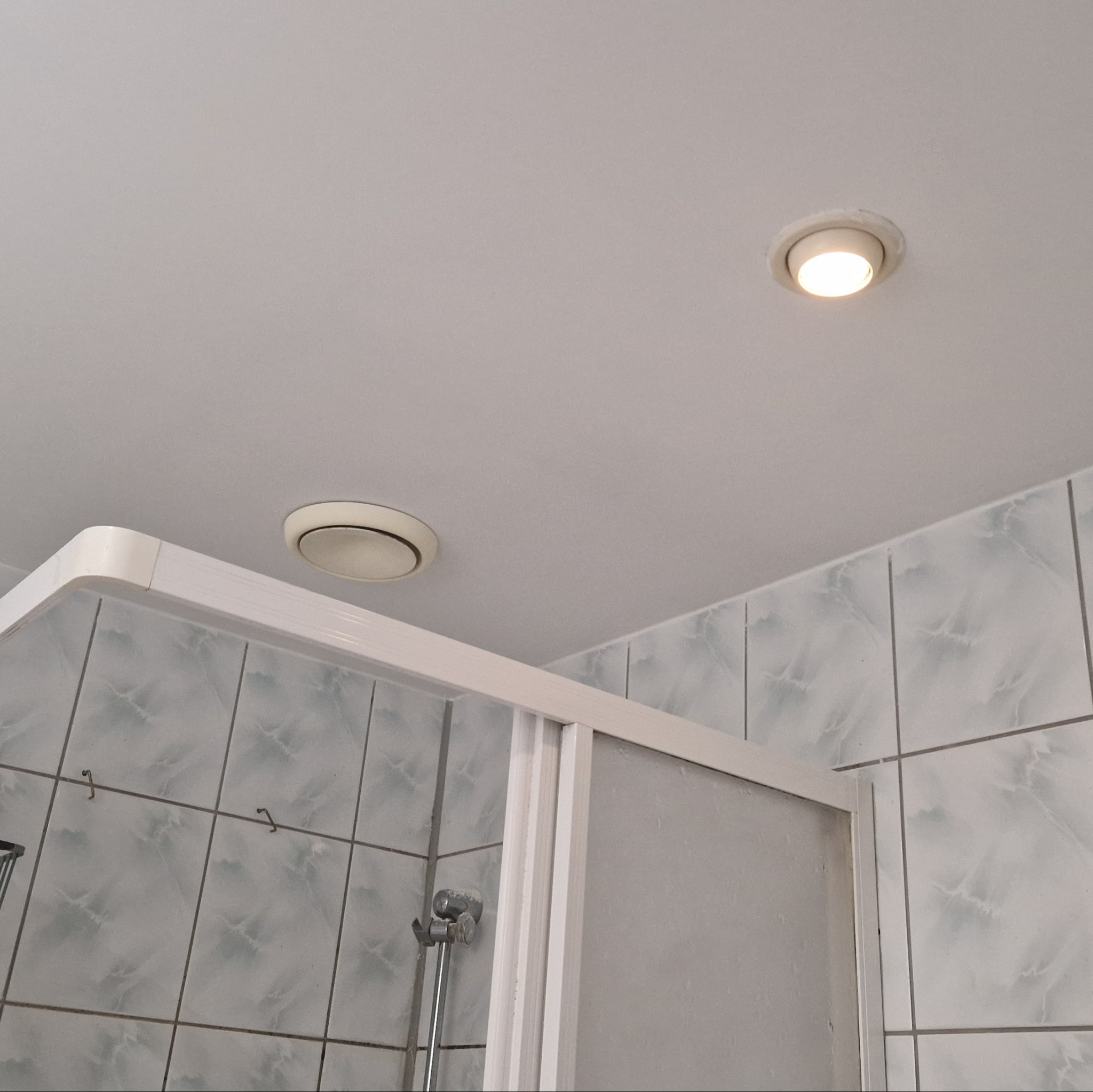 Fragment łazienki z kabiną prysznicową, płytkami w odcieniach szarości i bieli oraz dwoma okrągłymi lampami sufitowymi, z których jedna jest włączona.