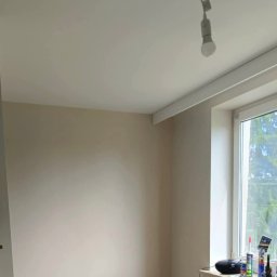Remontowo Sebastian Sandomierski - Jasne wnętrze po malowaniu ścian i sufitu, widoczne okno z widokiem na zieleń, lampa sufitowa i akcesoria malarskie na parapecie. Świeżo odnowione pomieszczenie.