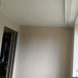 Remontowo Sebastian Sandomierski - Jasne wnętrze po remoncie: świeżo pomalowane ściany w odcieniach beżu, drewniana podłoga, listwy przypodłogowe i oświetlenie LED przy suficie. Widok na otwarte drzwi.