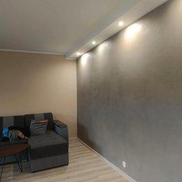 Remontowo Sebastian Sandomierski - Szara ściana z efektem dekoracyjnym, oświetlona punktowo, w salonie z szarą sofą i jasną podłogą. Widoczne wykończenie wnętrza.