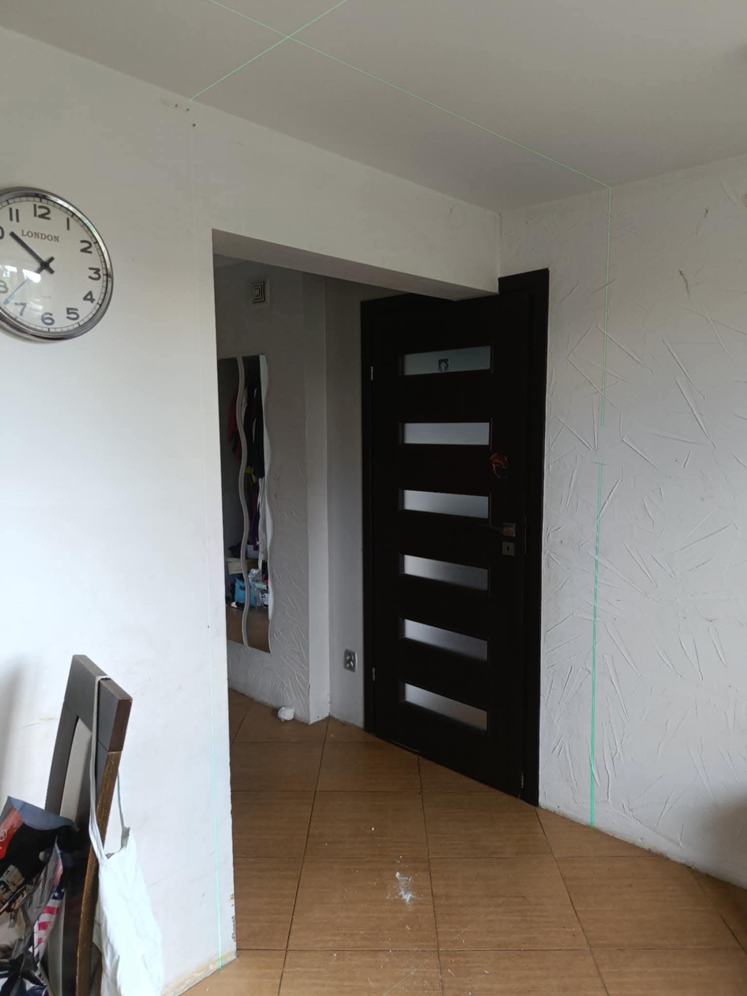 Korytarz w trakcie prac wykończeniowych: widoczne laserowe wyznaczniki na ścianie z fakturą, drzwi z przeszkleniami i zegar na ścianie. Ujęcie z poziomu podłogi.