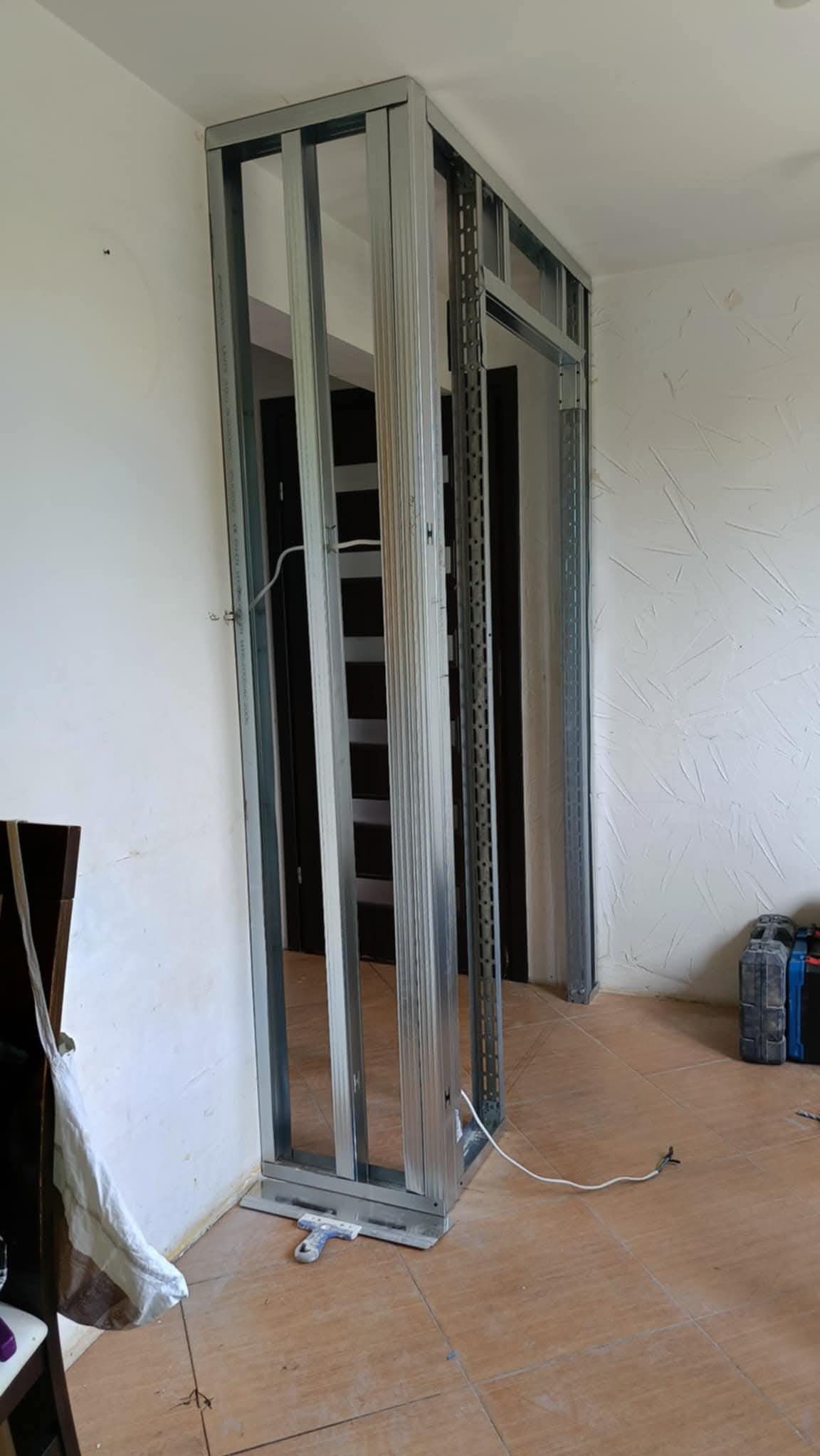 Metalowa konstrukcja ścianki działowej z profili, widoczna na tle białej ściany i brązowej podłogi. W tle widoczne drzwi i narzędzia.