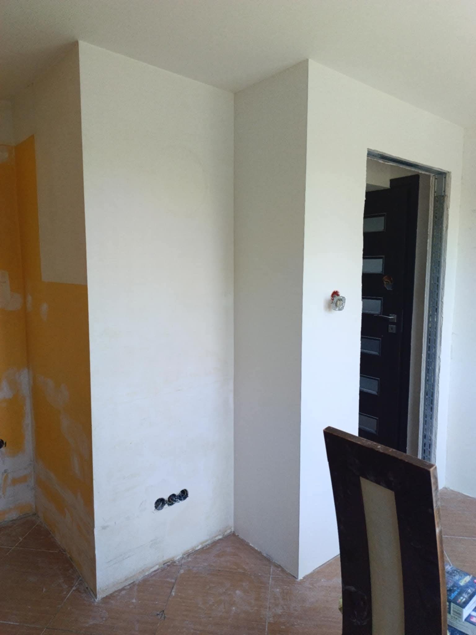 Fragment pokoju po gładziowaniu i malowaniu na biało, widoczne gniazdka elektryczne i fragment ściany w kolorze żółtym przed wykończeniem. Częściowo widoczne krzesło.