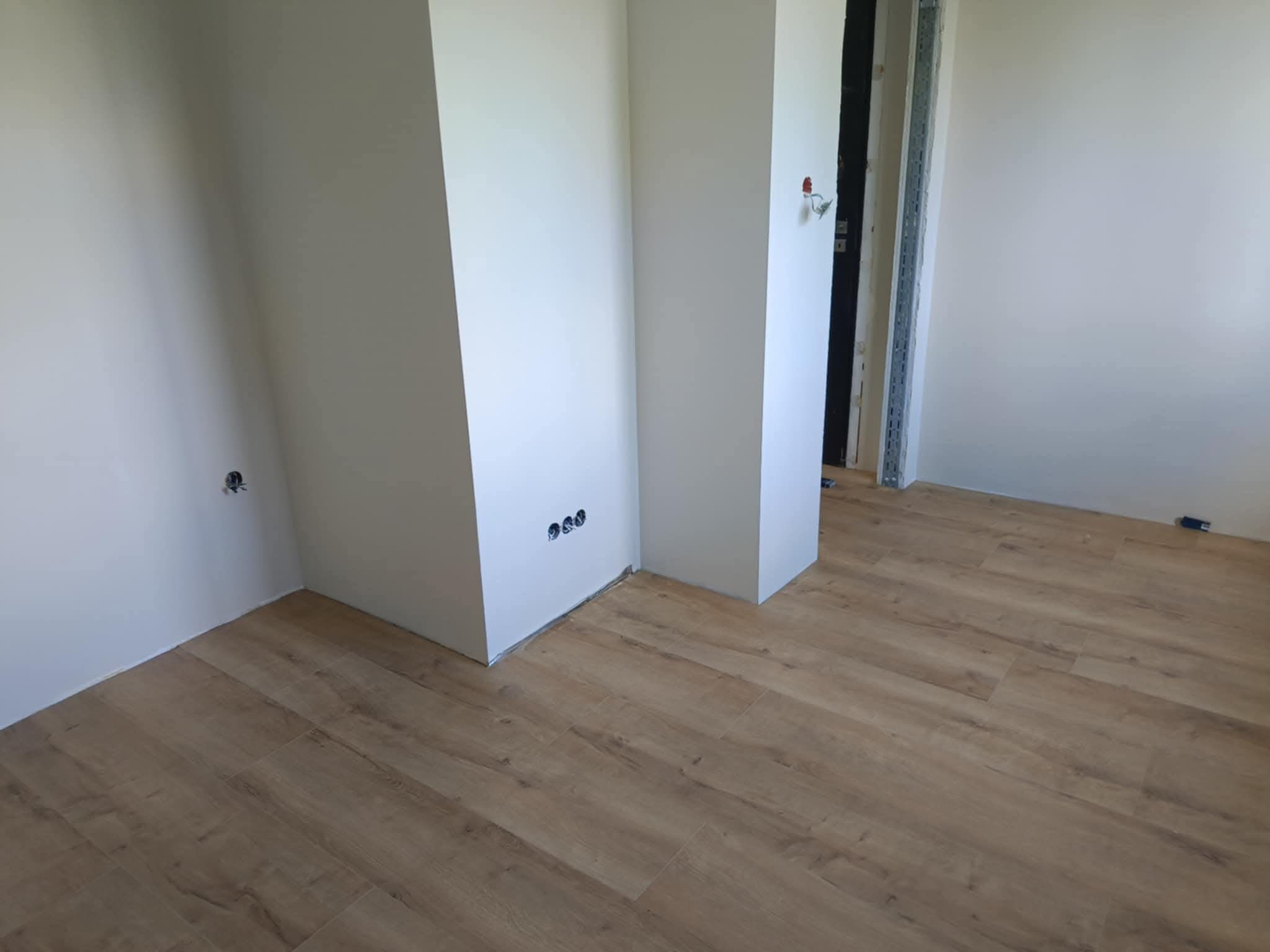 Jasne panele podłogowe w nowym wnętrzu z białymi ścianami i wystającymi elementami konstrukcyjnymi. Widoczne gniazdka elektryczne.