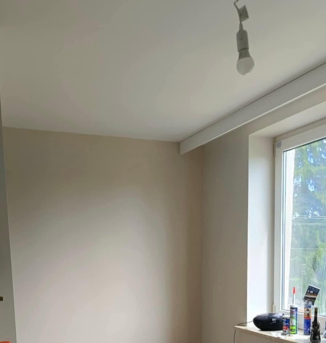 Jasne wnętrze po malowaniu ścian i sufitu, widoczne okno z widokiem na zieleń, lampa sufitowa i akcesoria malarskie na parapecie. Świeżo odnowione pomieszczenie.