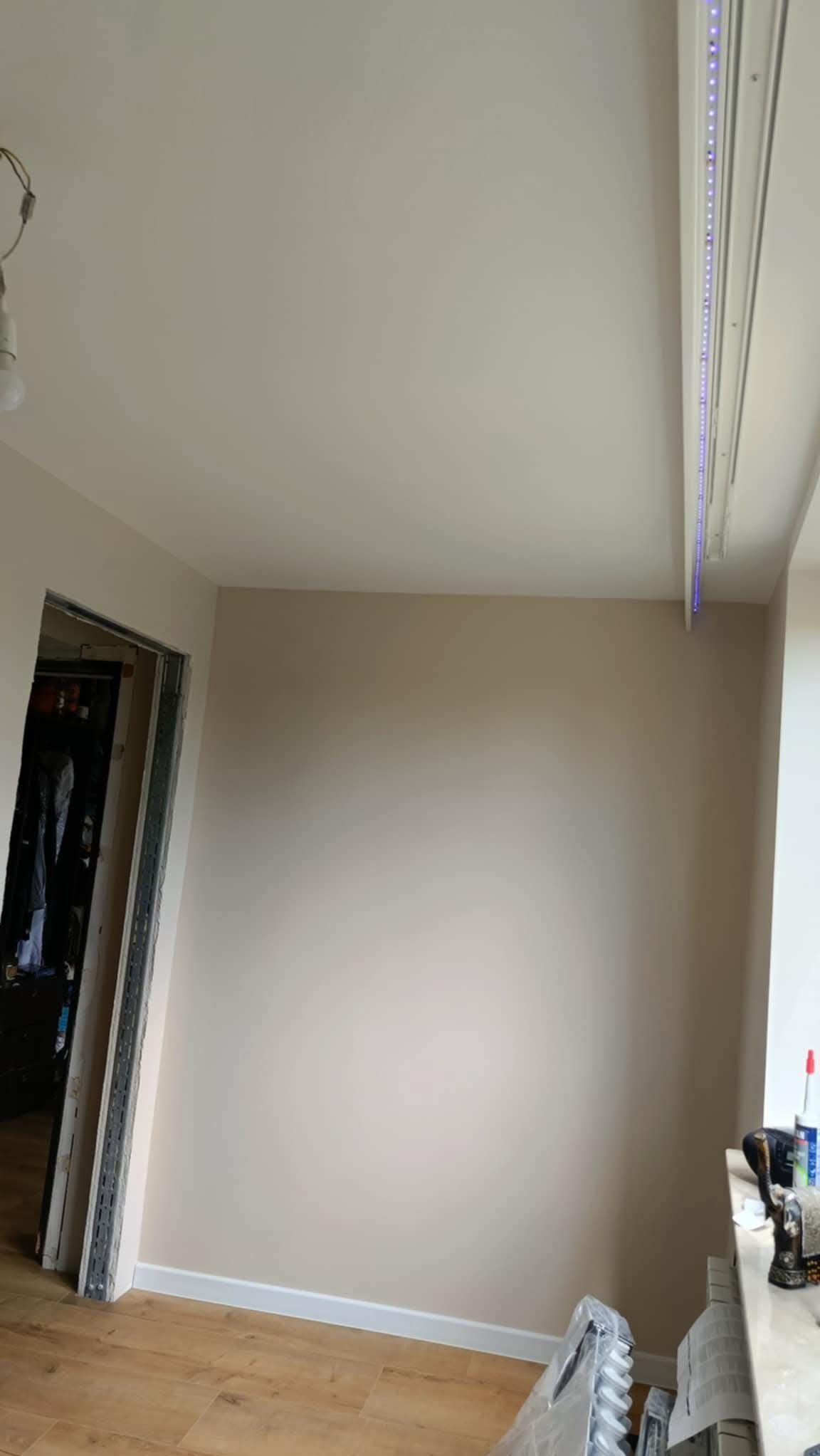 Jasne wnętrze po remoncie: świeżo pomalowane ściany w odcieniach beżu, drewniana podłoga, listwy przypodłogowe i oświetlenie LED przy suficie. Widok na otwarte drzwi.