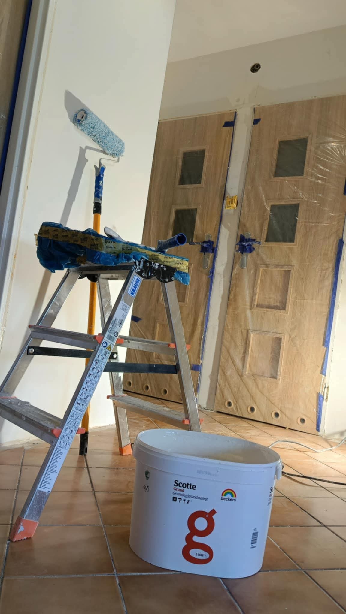 Przygotowania do malowania: drabina z wałkiem, pędzlami i wiadro farby Beckers Scotte na brązowej podłodze z płytek. Drzwi zabezpieczone folią malarską.