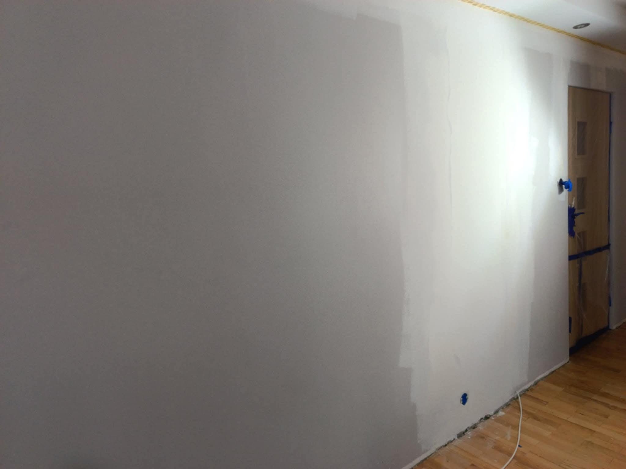 Ściana po szpachlowaniu, częściowo pomalowana na szaro, z widocznymi śladami taśmy malarskiej i drewnianą podłogą. Widoczny fragment drzwi zabezpieczonych folią.