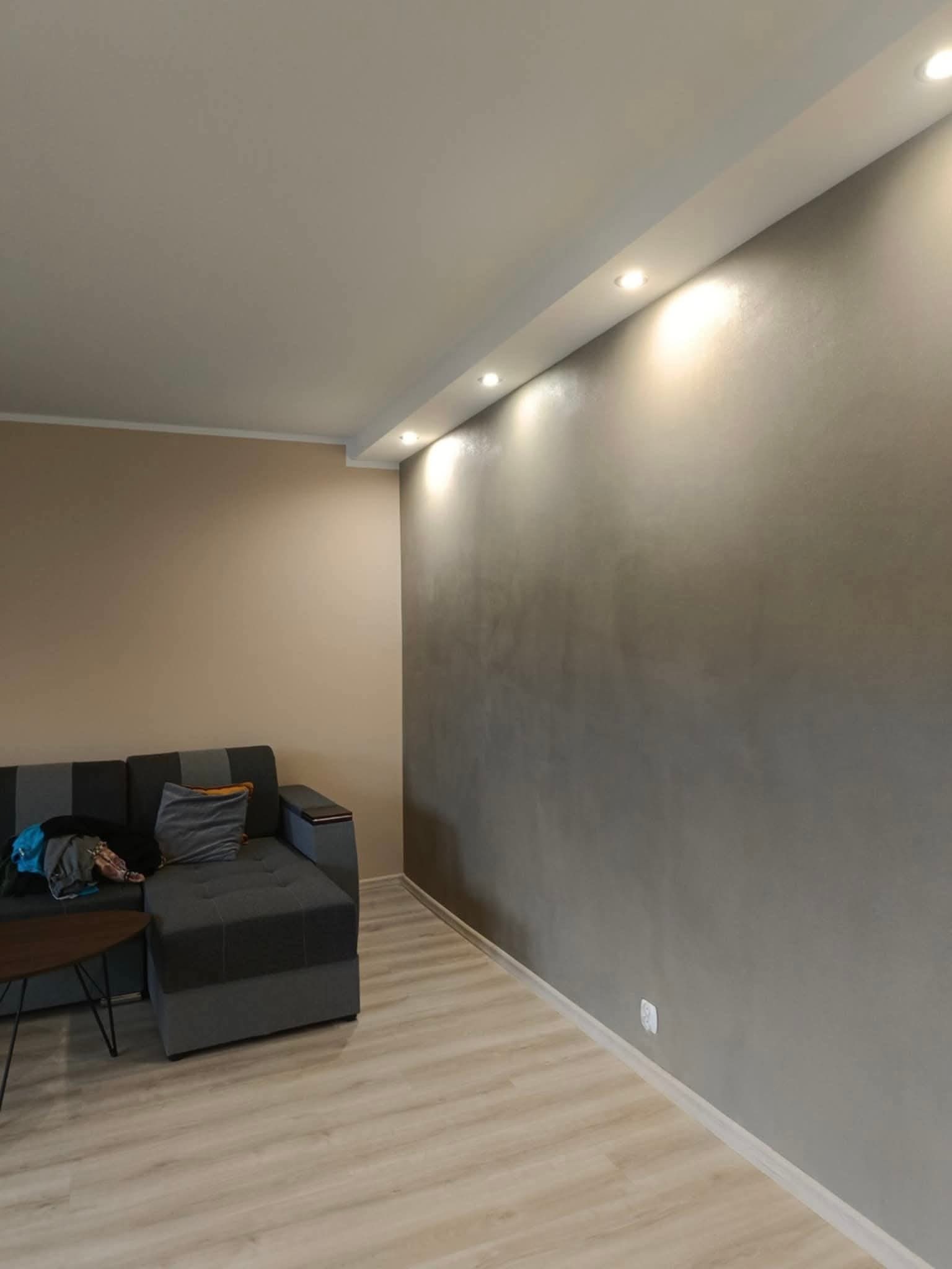 Szara ściana z efektem dekoracyjnym, oświetlona punktowo, w salonie z szarą sofą i jasną podłogą. Widoczne wykończenie wnętrza.