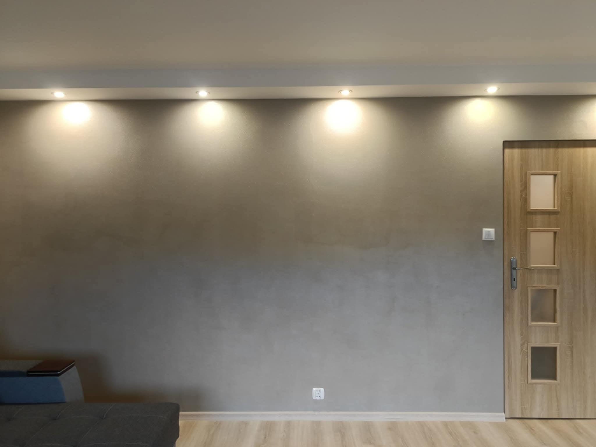 Szara ściana z efektem dekoracyjnym, oświetlona punktowo, z drewnianymi drzwiami po prawej stronie i fragmentem sofy w lewym dolnym rogu. Podłoga wyłożona panelami.