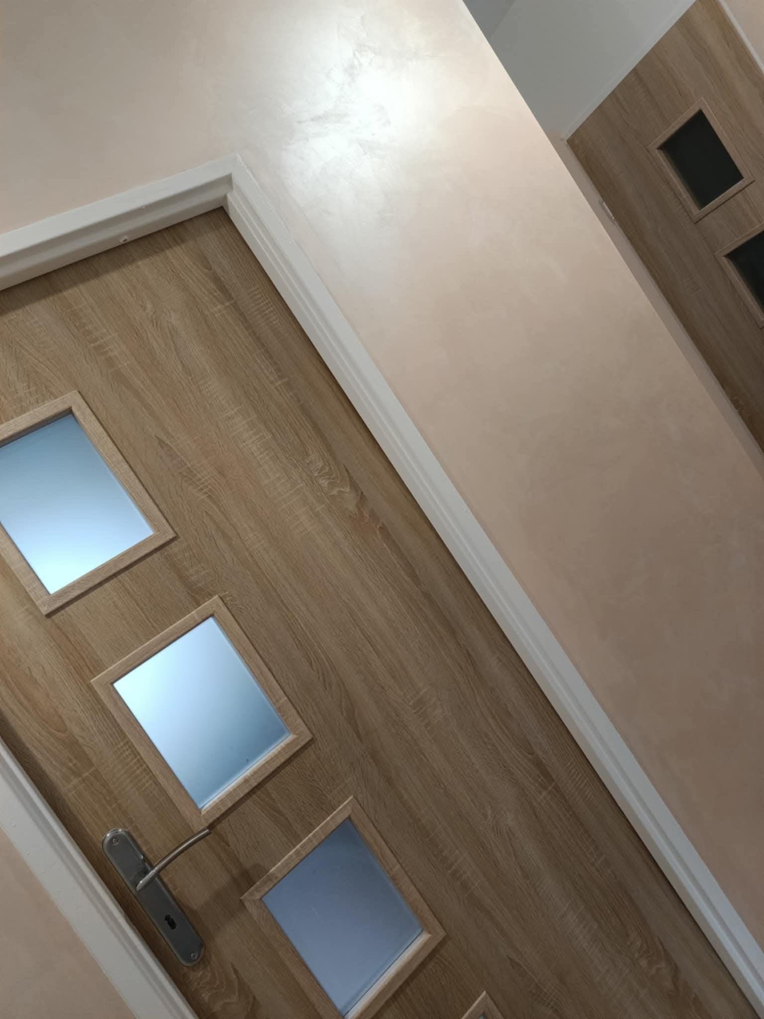 Beżowa ściana z efektem dekoracyjnym i drewniane drzwi z szybkami, wykończone białą listwą. Widok na fragment korytarza z drugimi drzwiami w tle.