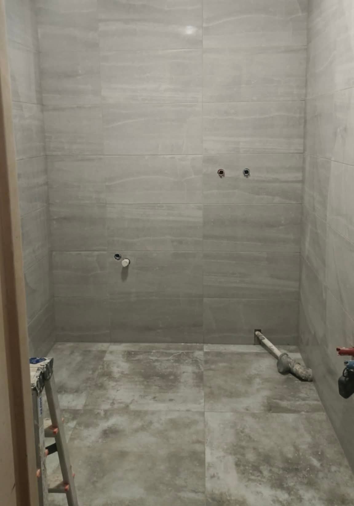 Wykończona łazienka z szarymi płytkami imitującymi kamień. Widoczne przyłącza wodne i kanalizacyjne. Na podłodze i ścianach ułożone płytki. Drabina malarska w rogu.