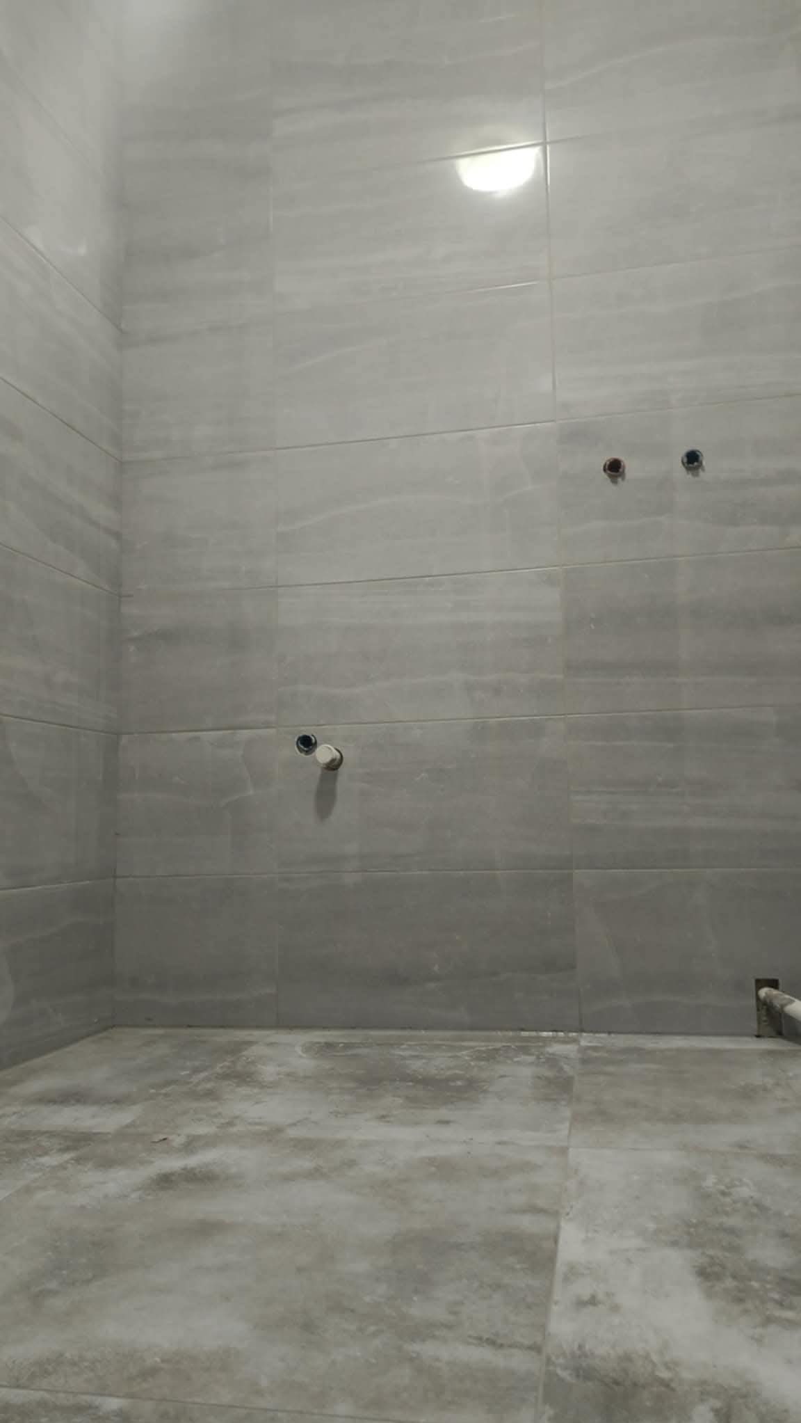 Szara łazienka z marmurowymi płytkami na ścianach i podłodze. Widoczne otwory na armaturę i rury. Minimalistyczny design, surowe wykończenie.