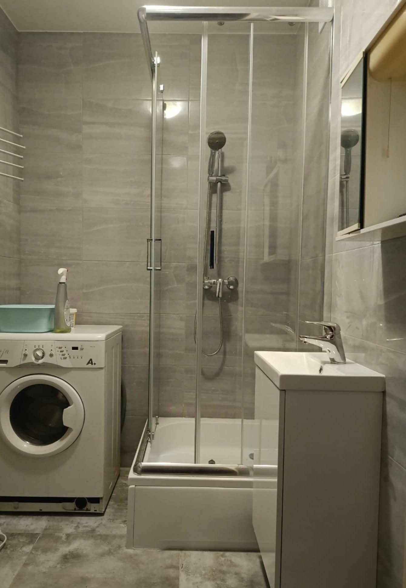 Wykończona łazienka w szarościach: kabina prysznicowa, umywalka z szafką, pralka. Minimalistyczny design, płytki imitujące kamień, chromowana armatura. Nowoczesne wnętrze.