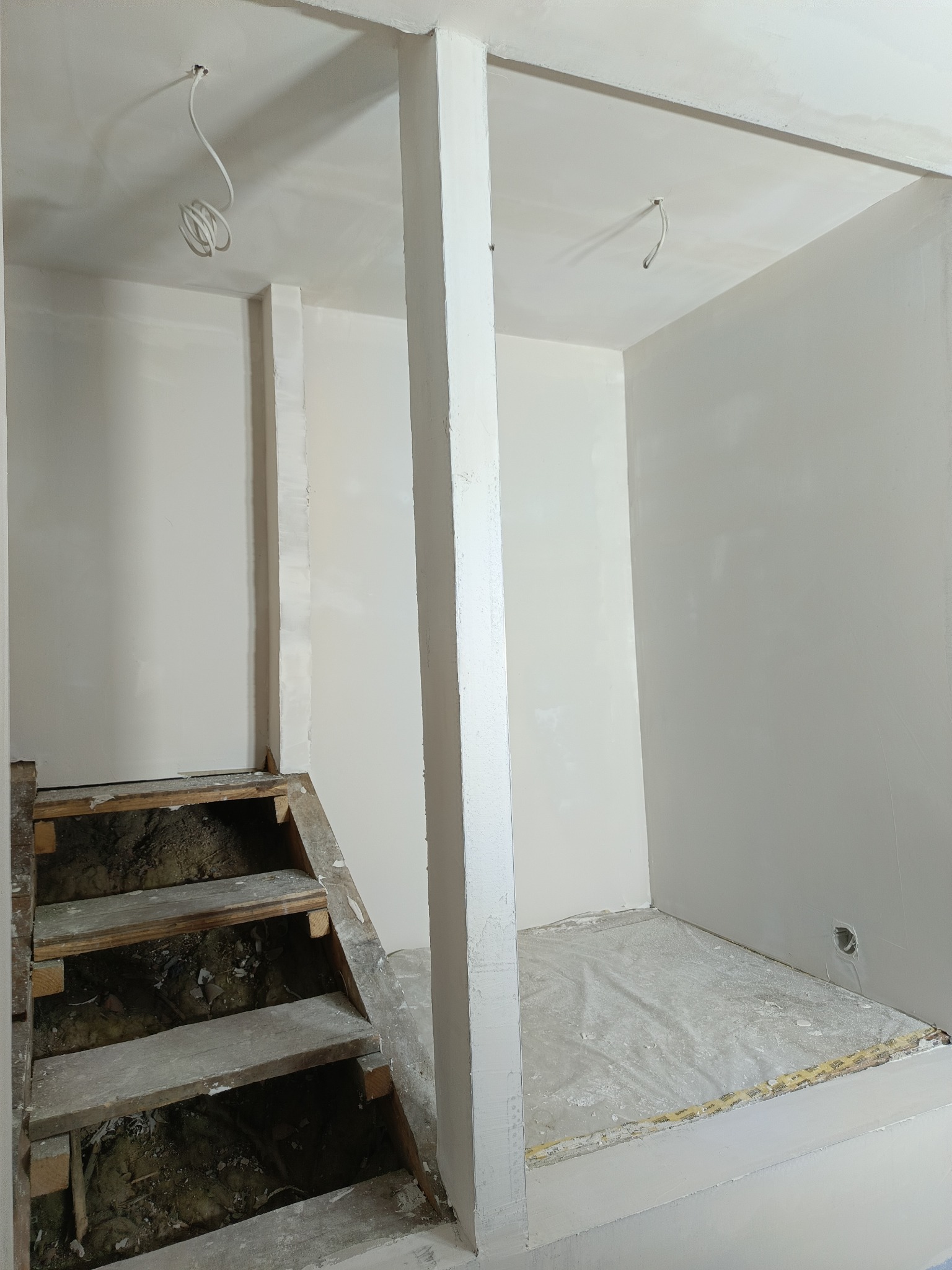 Wnętrze w trakcie prac wykończeniowych: gładkie, białe ściany, drewniane schody z widocznym podłożem i wystające kable elektryczne z sufitu.