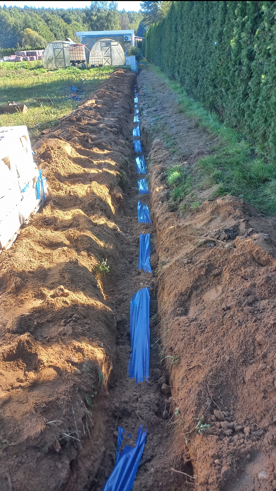 Wykop w ziemi z niebieską folią, prawdopodobnie przygotowany pod przyłącze elektryczne. Widoczne elementy infrastruktury w tle.