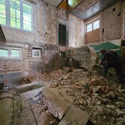 Market Group Sp. z o. o. - Demontaż ceglanej ściany wewnątrz budynku. Mężczyzna w roboczym ubraniu kuje ścianę, gruz leży na podłodze. Widoczny wózek do wywozu gruzu.