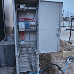 Market Group Sp. z o. o. - Otwarta skrzynka elektryczna z widocznymi bezpiecznikami i przewodami, umieszczona na zewnątrz budynku, częściowo zagłębiona w ziemi. Widoczne narzędzia i elementy konstrukcyjne.