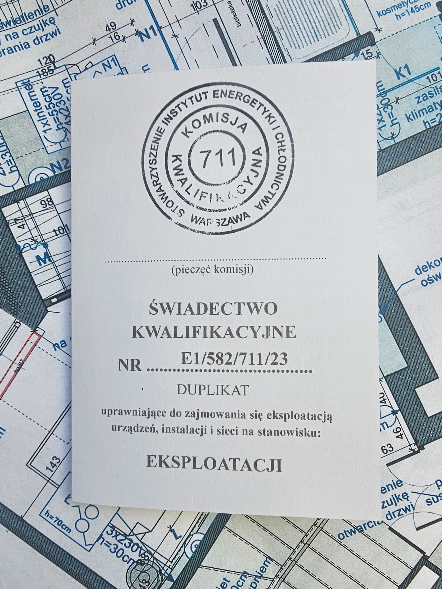 Duplikat świadectwa kwalifikacyjnego E1/582/711/23 na tle planu instalacji elektrycznej budynku.