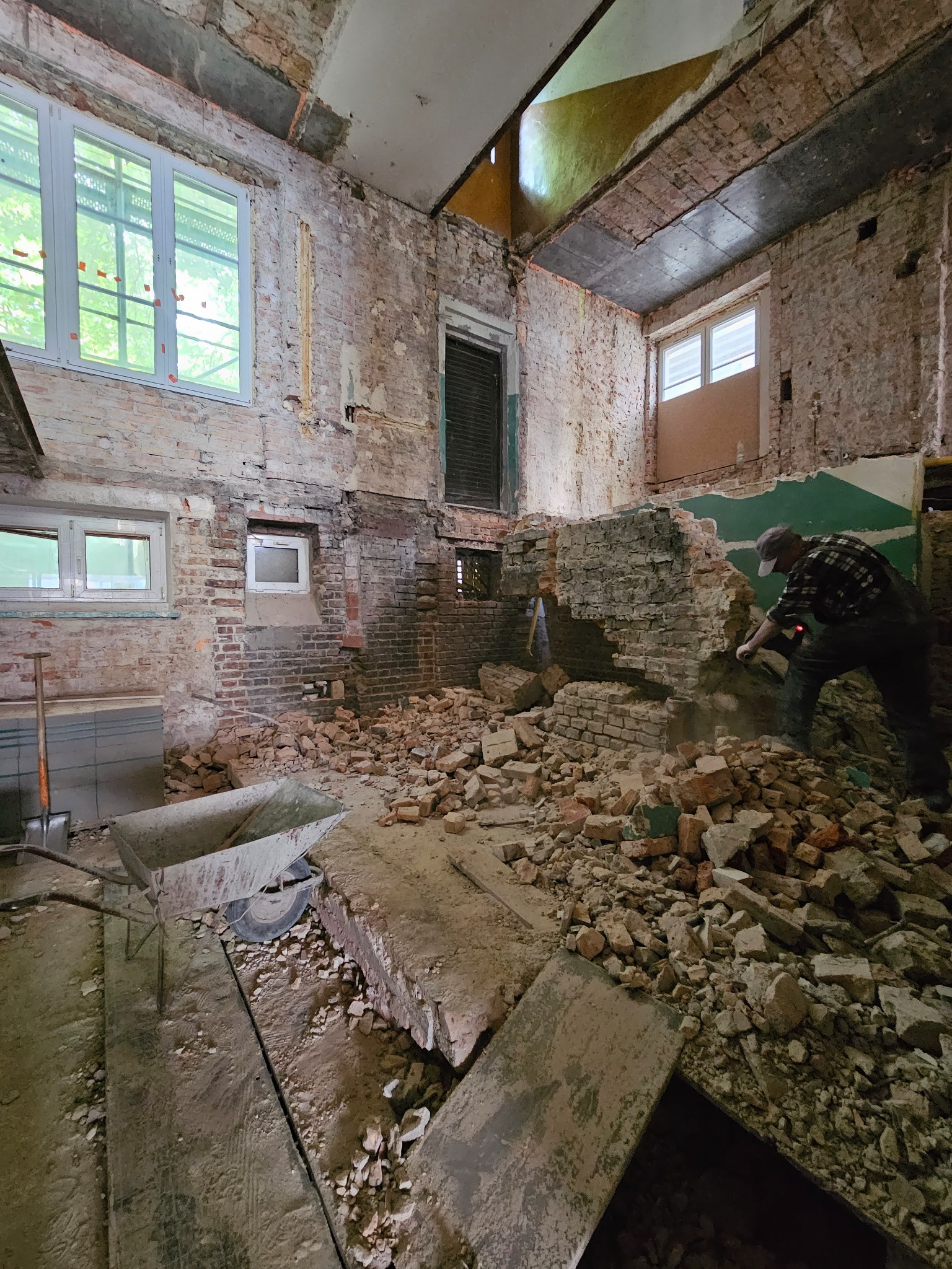 Demontaż ceglanej ściany wewnątrz budynku. Mężczyzna w roboczym ubraniu kuje ścianę, gruz leży na podłodze. Widoczny wózek do wywozu gruzu.