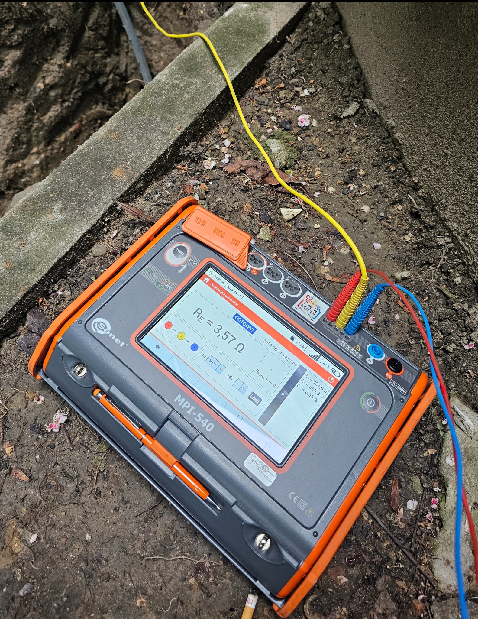 Miernik Sonel MPI-540 z podłączonymi przewodami pomiarowymi, leżący na ziemi, wskazuje rezystancję uziemienia 3,57 Ω. Widoczne fragmenty betonu i ziemi w tle.