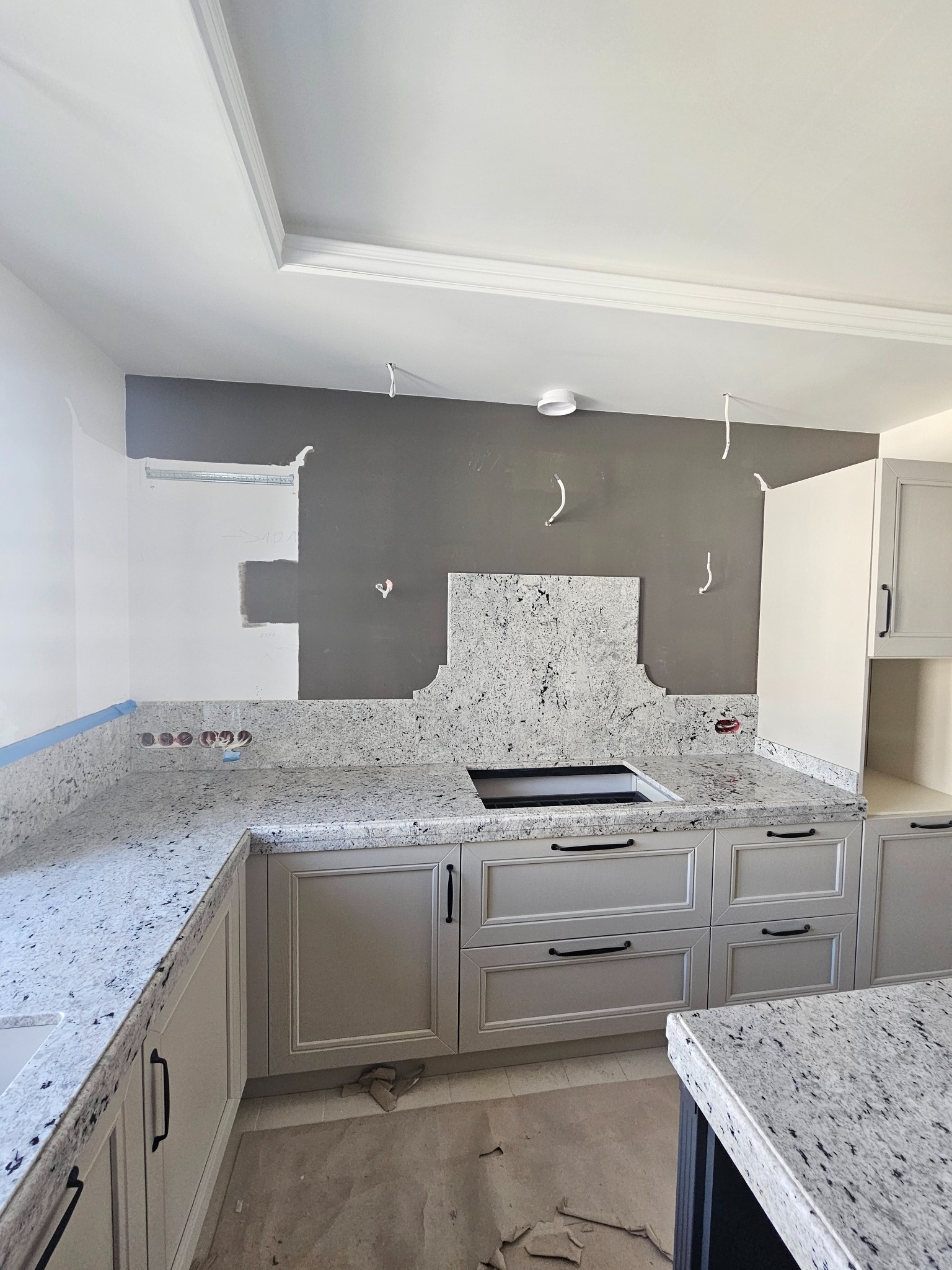 Kuchnia w trakcie montażu: szare szafki z granitowym blatem, widoczne kable i otwory na gniazdka, fragmenty pomalowanej ściany, sufit z dekoracyjną listwą.