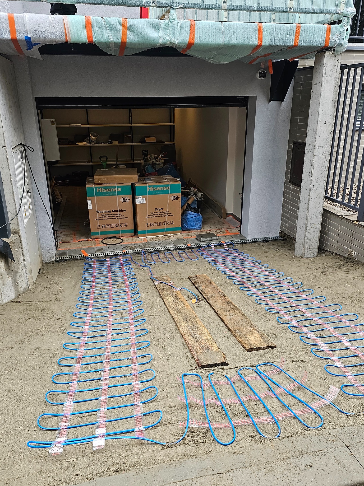 Instalacja ogrzewania podjazdowego z niebieskich rurek na piasku przed garażem. Widoczne kartony po pralkach i suszarkach. Ujęcie z góry, szeroki kadr.