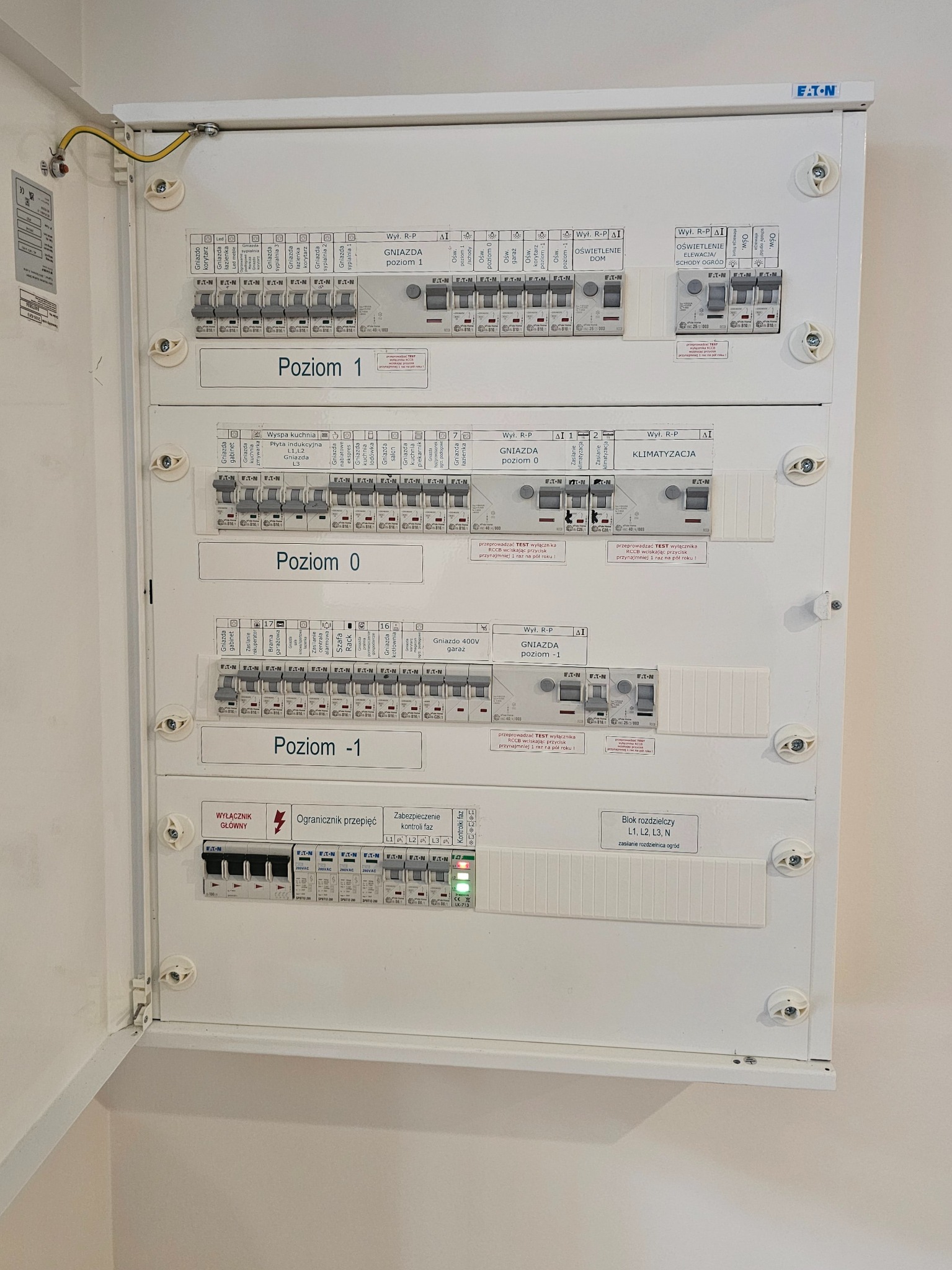 Otwarta, biała szafka rozdzielcza Eaton z bezpiecznikami i oznaczeniami poziomów (1, 0, -1) oraz obwodów (gniazda, oświetlenie, klimatyzacja). Widoczne zabezpieczenia i blok rozdzielczy.