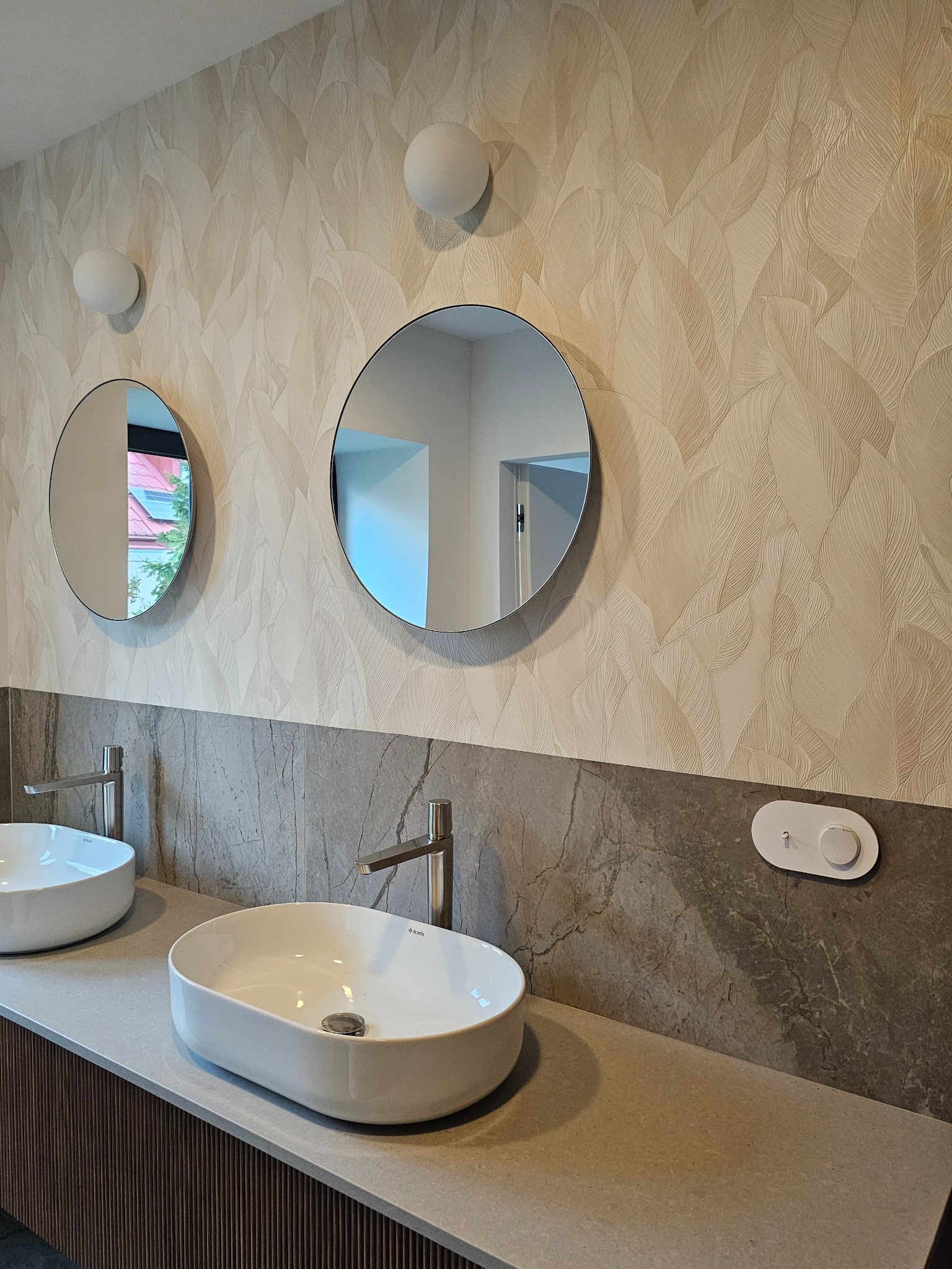 Nowoczesna łazienka z dwoma okrągłymi umywalkami i lustrami, oświetlona kulistymi lampami na tle ściany w liściasty wzór. Widoczny panel sterowania.