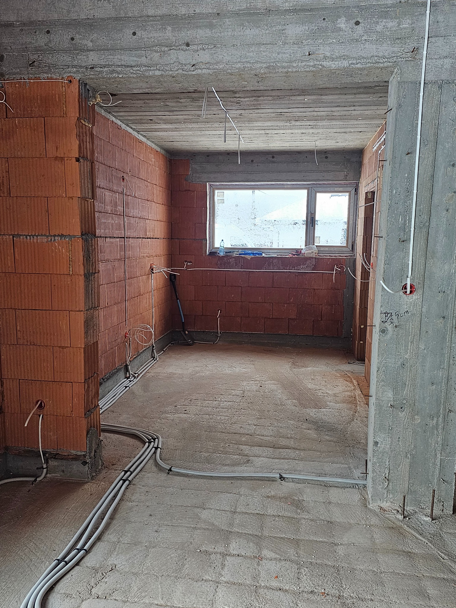 Surowy stan wnętrza z widoczną instalacją elektryczną: kable w peszlach ułożone na podłodze, wyprowadzenia ze ściany, puszka elektryczna na słupie. Cegła dziurawka.