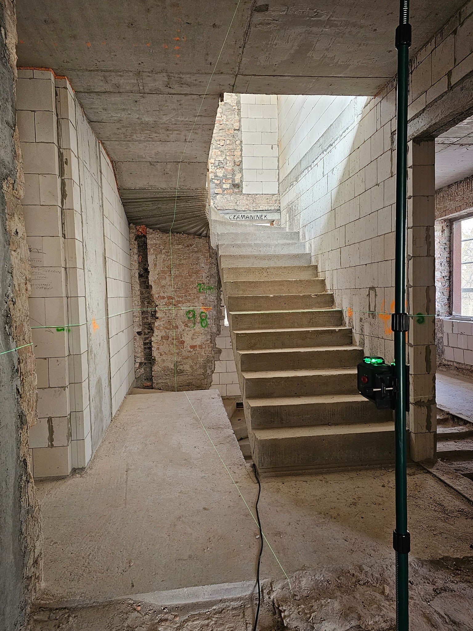 Wnętrze w trakcie remontu: surowe ściany z betonu komórkowego i cegły, betonowe schody, zielony laser krzyżowy wyznaczający poziom. Widoczne ślady po instalacjach.