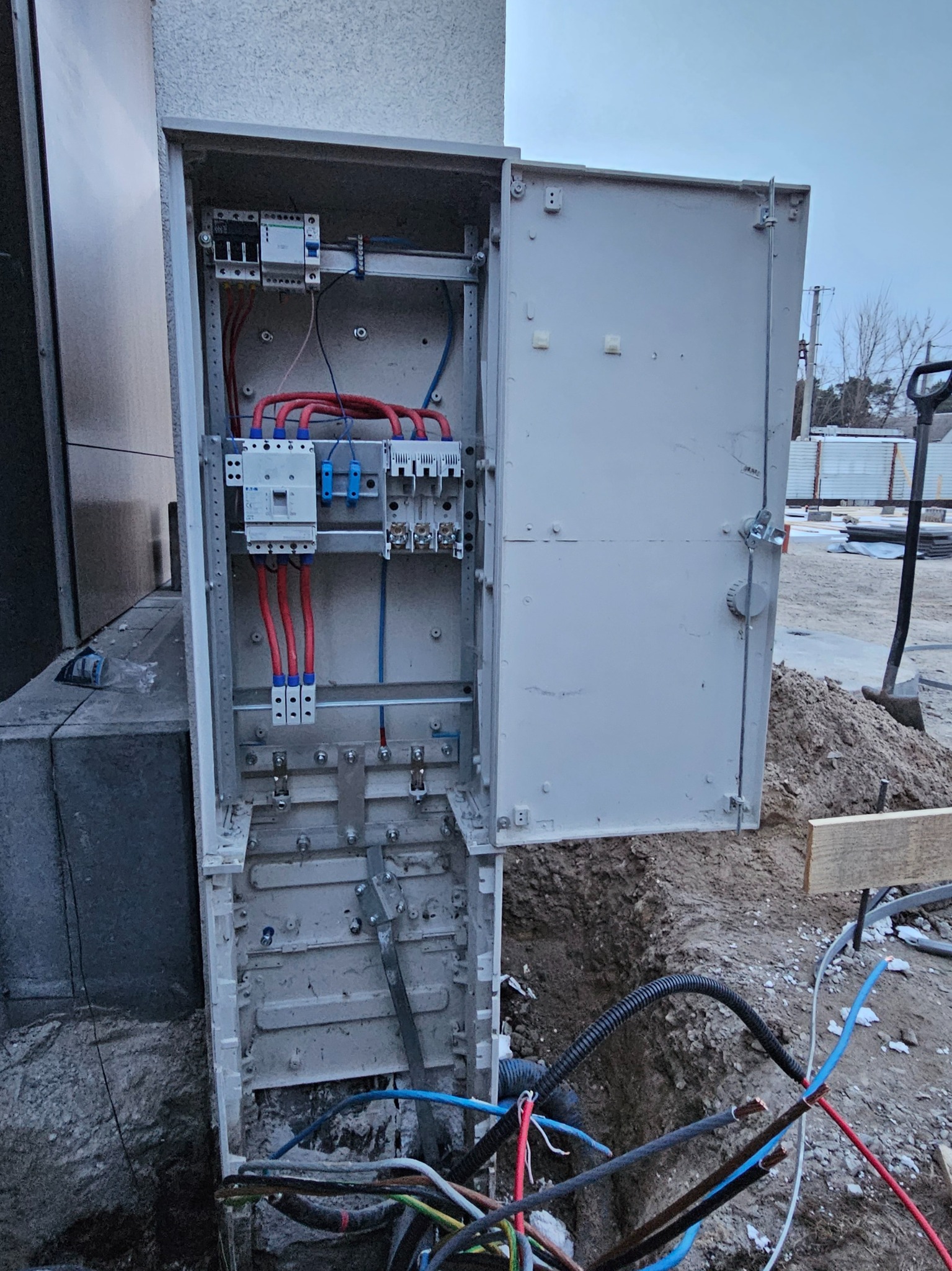 Otwarta skrzynka elektryczna z widocznymi bezpiecznikami i przewodami, umieszczona na zewnątrz budynku, częściowo zagłębiona w ziemi. Widoczne narzędzia i elementy konstrukcyjne.