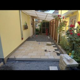 Jan Chmielarz - Układanie jasnych płyt chodnikowych na podsypce żwirowej, widoczna korytka odpływowe, w tle ściana budynku w kolorze żółtym i drewniana konstrukcja z zadaszeniem z poliwęglanu.