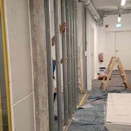 Jan Chmielarz - Montaż metalowego stelażu do ścian działowych w biurze, widoczna instalacja wentylacyjna pod sufitem, drabina i materiały budowlane. Na podłodze rozłożona mata ochronna.