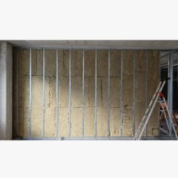 Jan Chmielarz - Ściana z metalowym stelażem wypełnionym wełną mineralną, widoczna drabina oparta o ścianę, etap prac wykończeniowych.