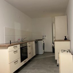 Jan Chmielarz - Wnętrze kuchni w trakcie prac wykończeniowych: położone szare płytki podłogowe, białe szafki kuchenne z drewnianym blatem, widoczna pralka i sprzęt AGD.