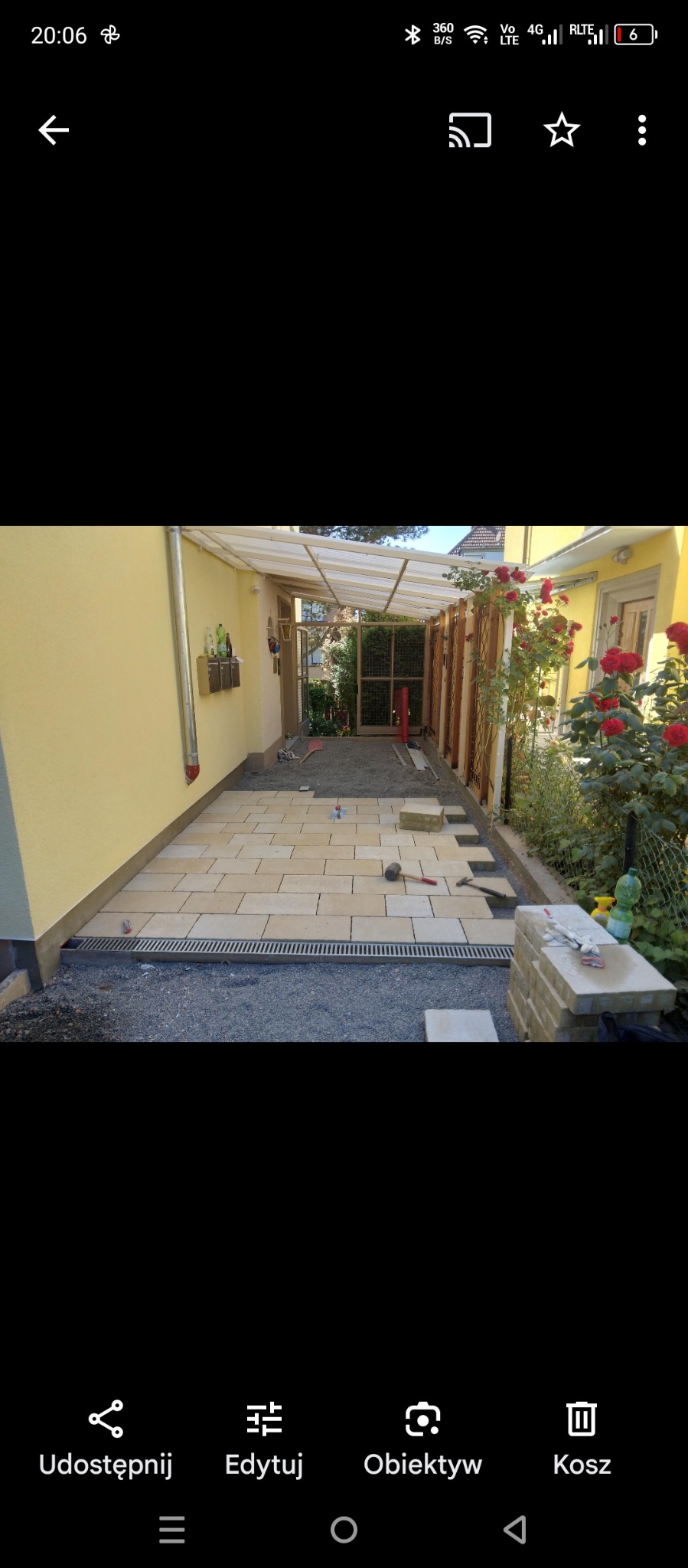 Układanie jasnych płyt chodnikowych na podsypce żwirowej, widoczna korytka odpływowe, w tle ściana budynku w kolorze żółtym i drewniana konstrukcja z zadaszeniem z poliwęglanu.