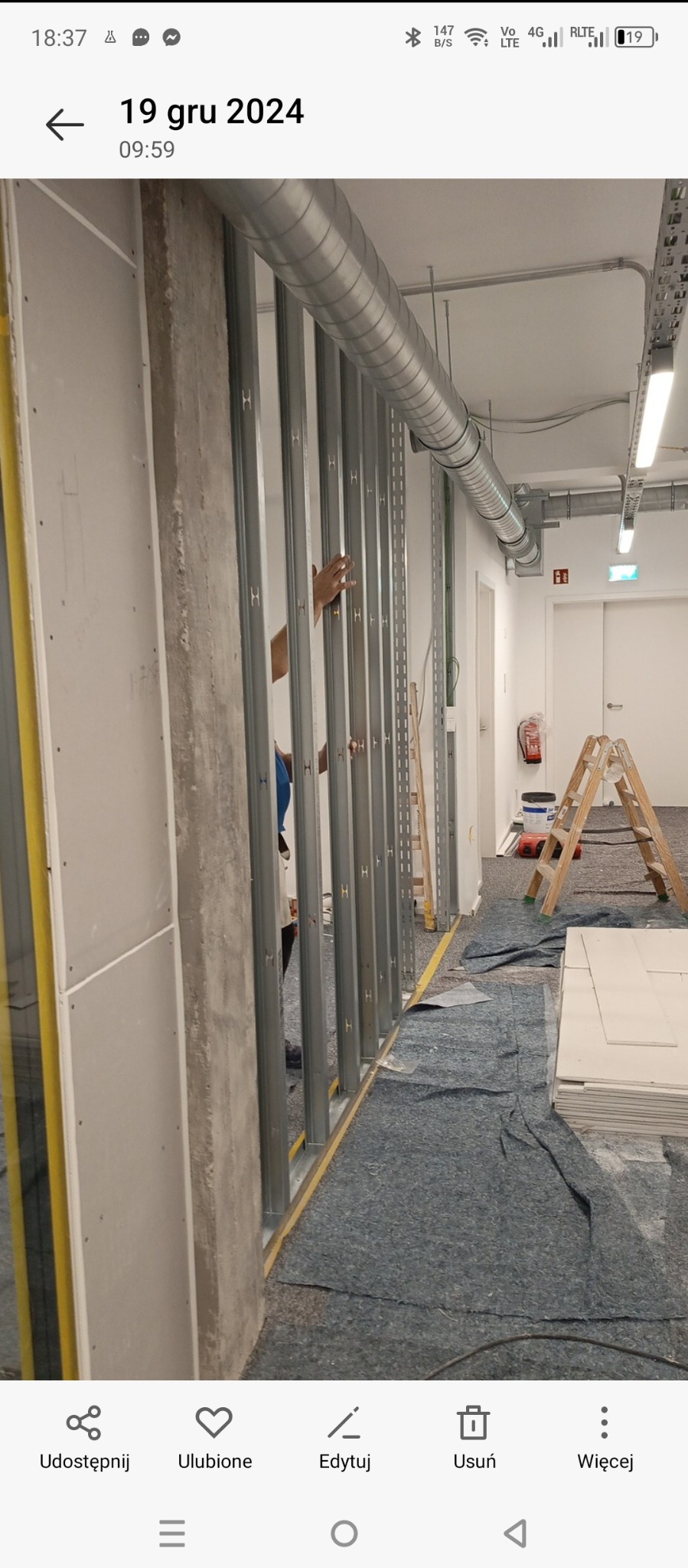 Montaż metalowego stelażu do ścian działowych w biurze, widoczna instalacja wentylacyjna pod sufitem, drabina i materiały budowlane. Na podłodze rozłożona mata ochronna.