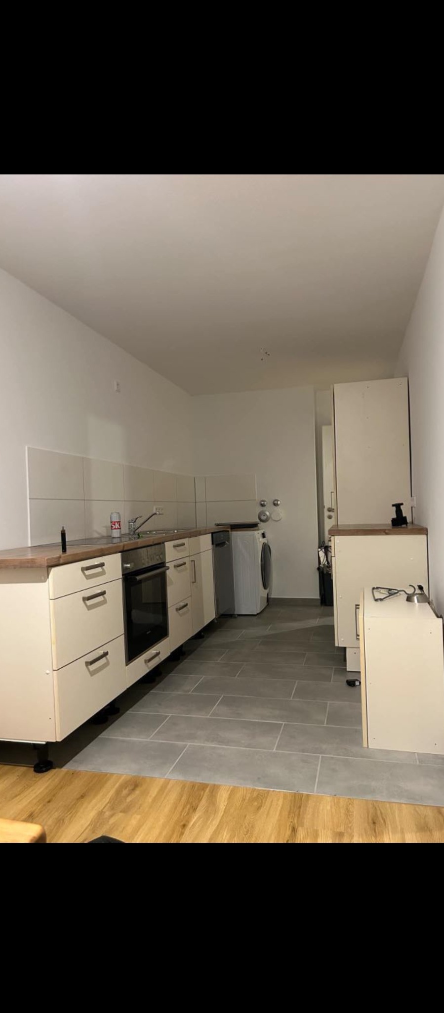 Wnętrze kuchni w trakcie prac wykończeniowych: położone szare płytki podłogowe, białe szafki kuchenne z drewnianym blatem, widoczna pralka i sprzęt AGD.