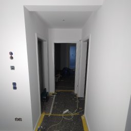 USŁUGI BUDOWLANE Oktawian Rożenko - Korytarz w trakcie malowania na biało, zabezpieczona podłoga, widoczne puszki elektryczne bez osprzętu, butelka wody i narzędzie elektryczne.